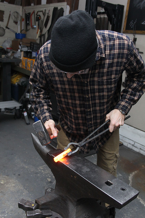 Blacksmith Tait Lawrence shaping hot steel using a hammer, tongs, and an anvil in his workshop.