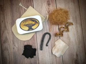 Looking down at the materials that make up a Raven's Roost Forge Flint Striker Kit. Materials include metal box with company logo, woven bag, char cloth, flint, kindling, and steel striker.