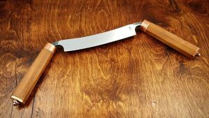 Looking down at a woodworkers draw knife with rounded handles, brass accents and a handmade steel blade.