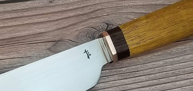 A close up of the “TL” touchmark with the “T” in the form of a cutlass, stamped into the blade of a steel knife with a pale wooden handle.